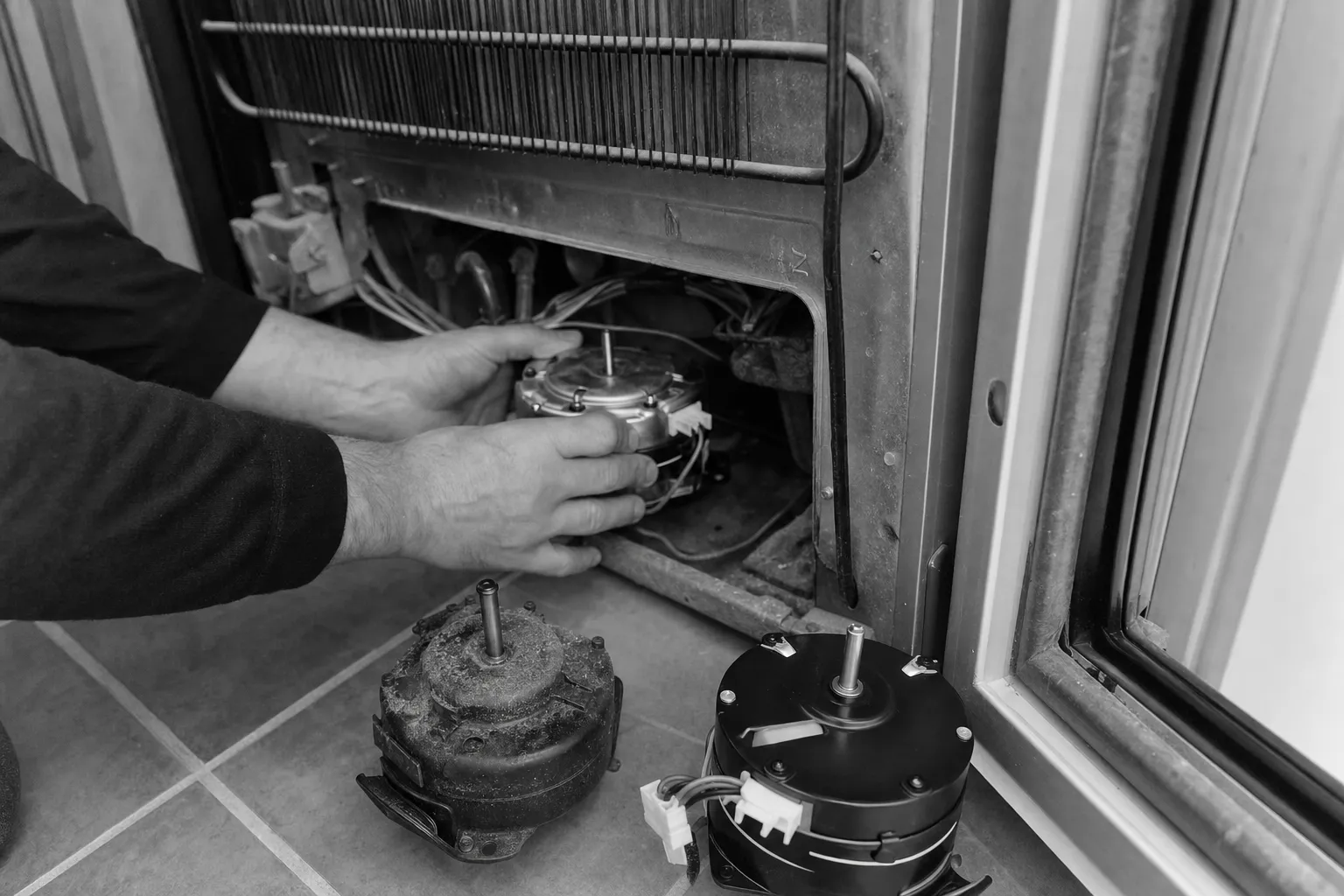Black-and-white frame of hands fitting a motor below a wire condenser, the old and new motors standing on a tiled floor